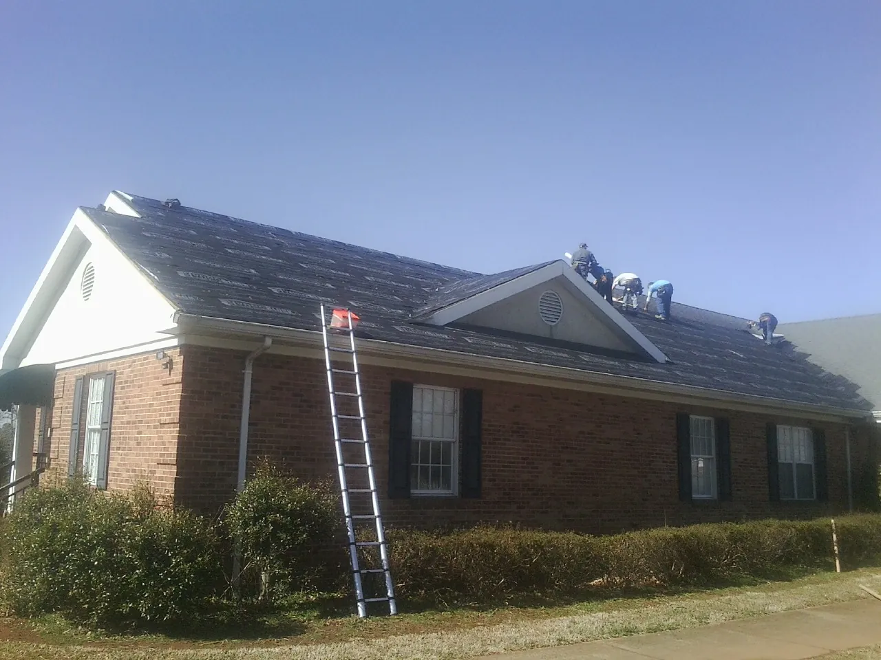 Crew tearing shingles off a brick ranch roof with a ladder set — Emergency Roof Repair work by our crew in Spirit Lake