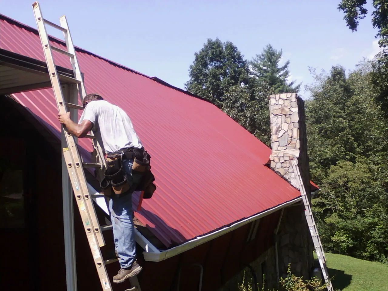 Roofer on a ladder beside a red metal roof and stone chimney — Soffit & Fascia Repair by Roofing Brothers in Spirit Lake
